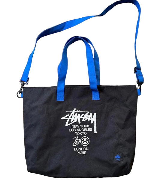 Black tote bag with blue straps and Stussy branding on a patterned rug